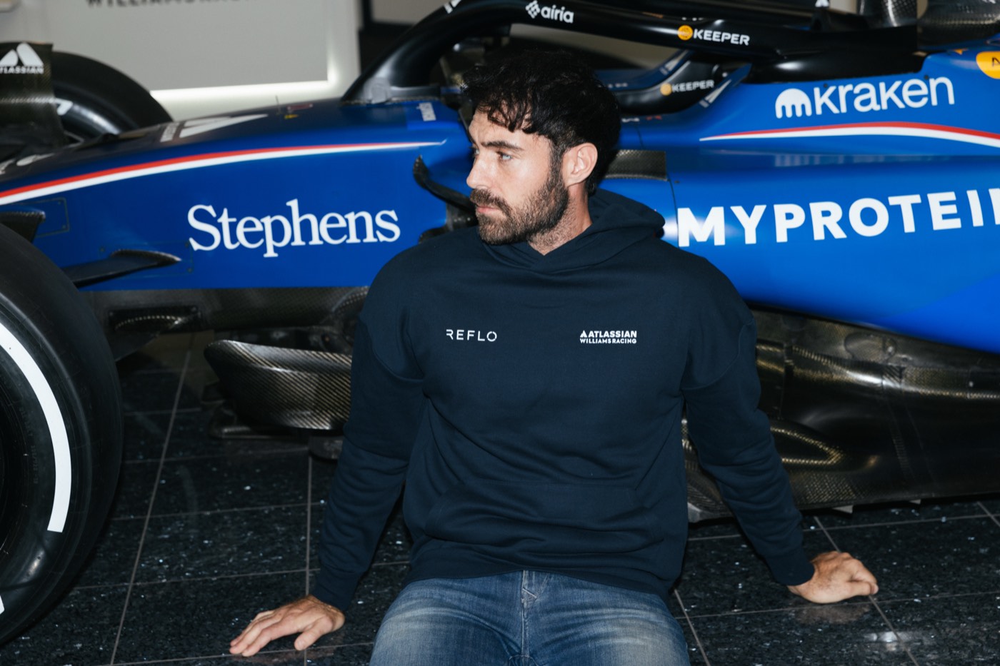 Crew member in a navy Reflo hoodie sitting on the garage floor in front of the Formula 1 car